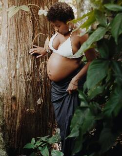 Black Pregnant Person posing against tree holding their baby belly.